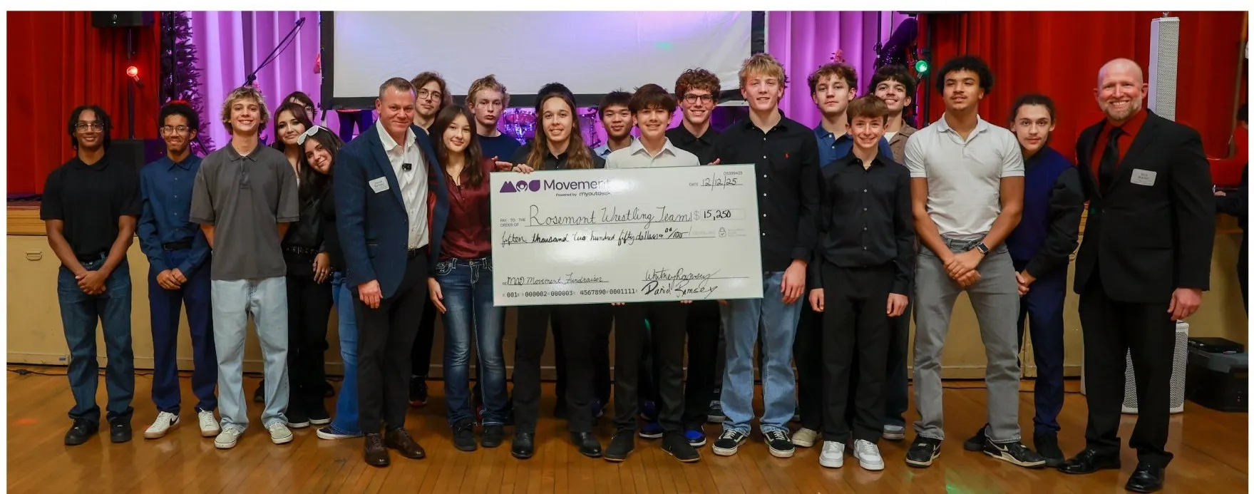 The Rosemont High School wrestling team on stage with Daniel Ramsey holding a large donation check at the 2025 Christmas Fundraiser